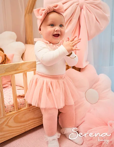 Bebê de saia de tule rosa em pé ao lado de um berço, marca Serena Baby.